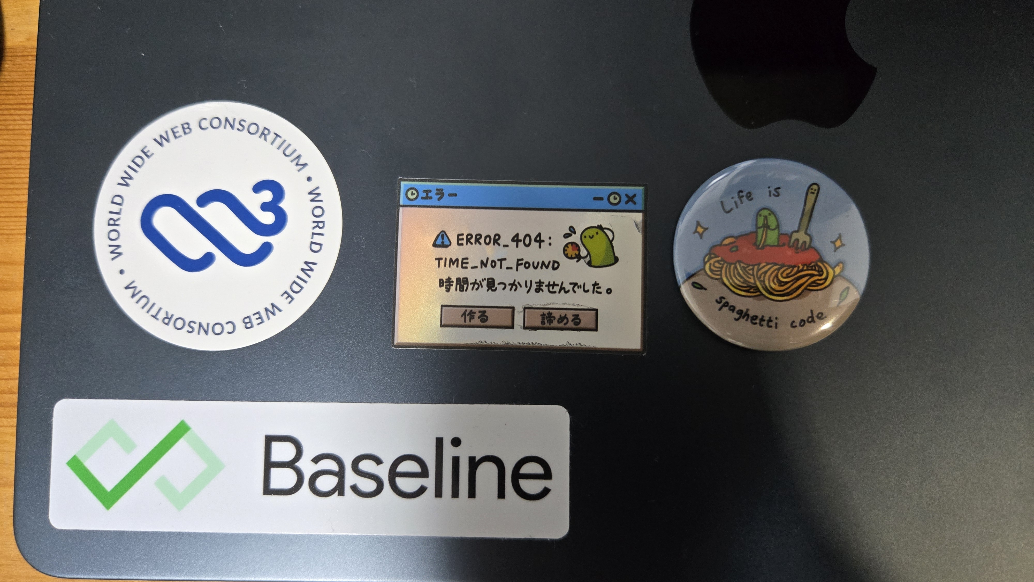 MacBookに入手したステッカーを貼り付けている。W3Cの新しいロゴのステッカー、バグちゃんの404 TIME_NOT_FOUNDのステッカー、スパゲッティコードならぬ本当にスパゲティを食べているバグちゃんの缶バッジ、Baselineのロゴステッカーが並んでいる